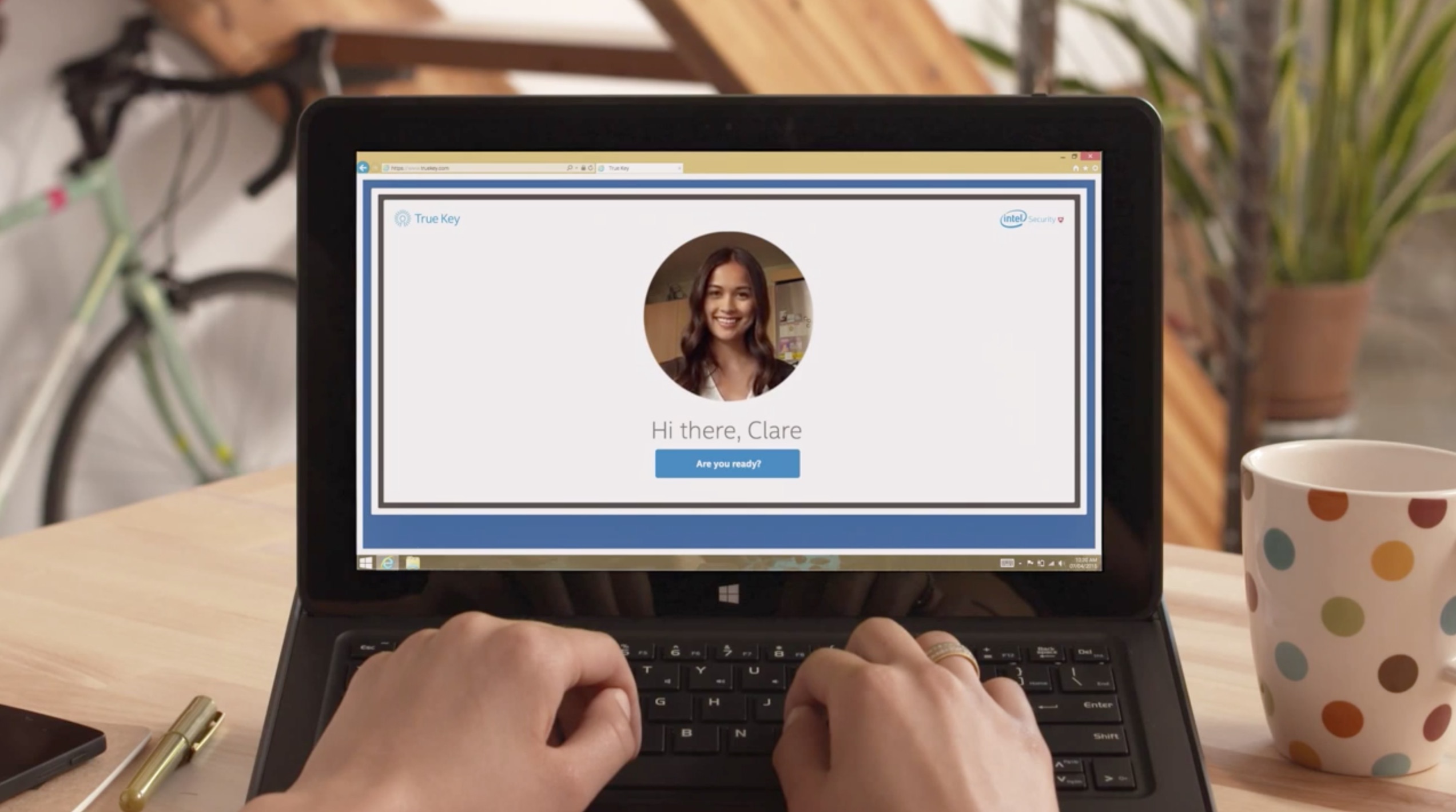 Hands typing at a laptop on a sunlit desk; the browser shows True Key recognising the person from her photo and greeting her — “Hi there, Clare”.