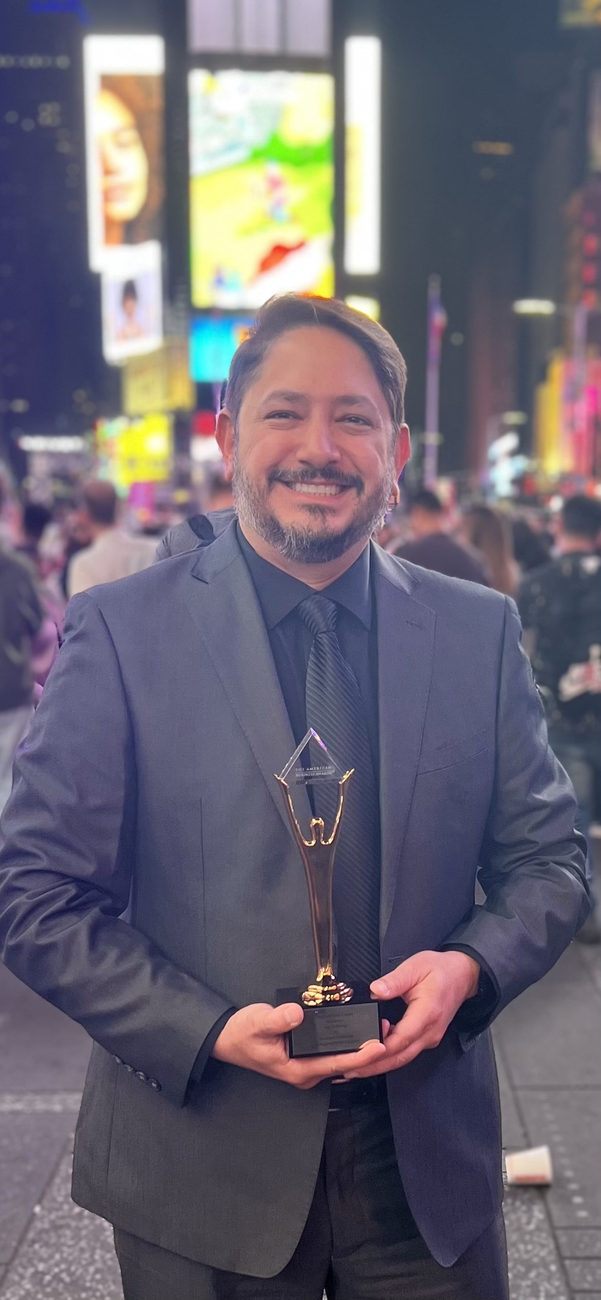 Marcus Couto standing in Times Square at night, holding the Gold Stevie Award trophy, with the lit billboards behind him.